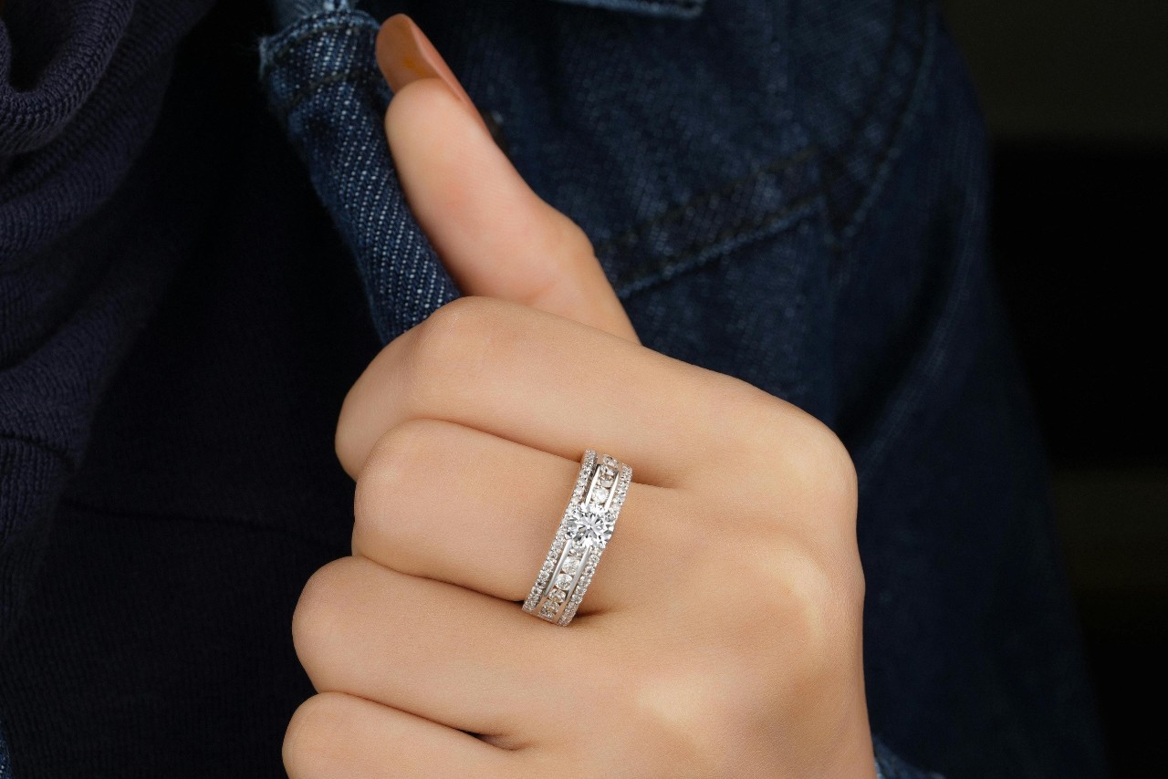 A person wearing a sparkling diamond band ring on their finger with a denim jacket visible in the background. A person wearing a sparkling diamond band ring on their finger with a denim jacket visible in the background.